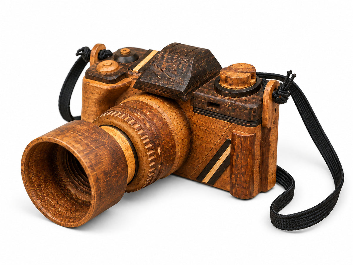 Handmade wooden camera sculpture viewed from the right side, showing its carved lens, wood grain, top controls and black strap against a light background.